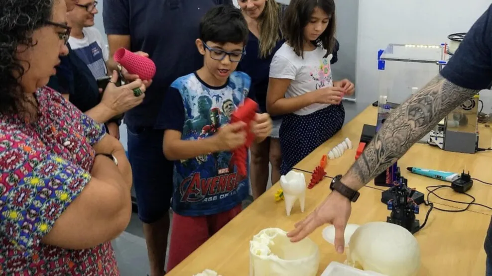 Crianças e adultos observam e seguram réplicas de artefatos impressas em 3D, como dentes e crânios, sobre uma mesa do Fab Lab.