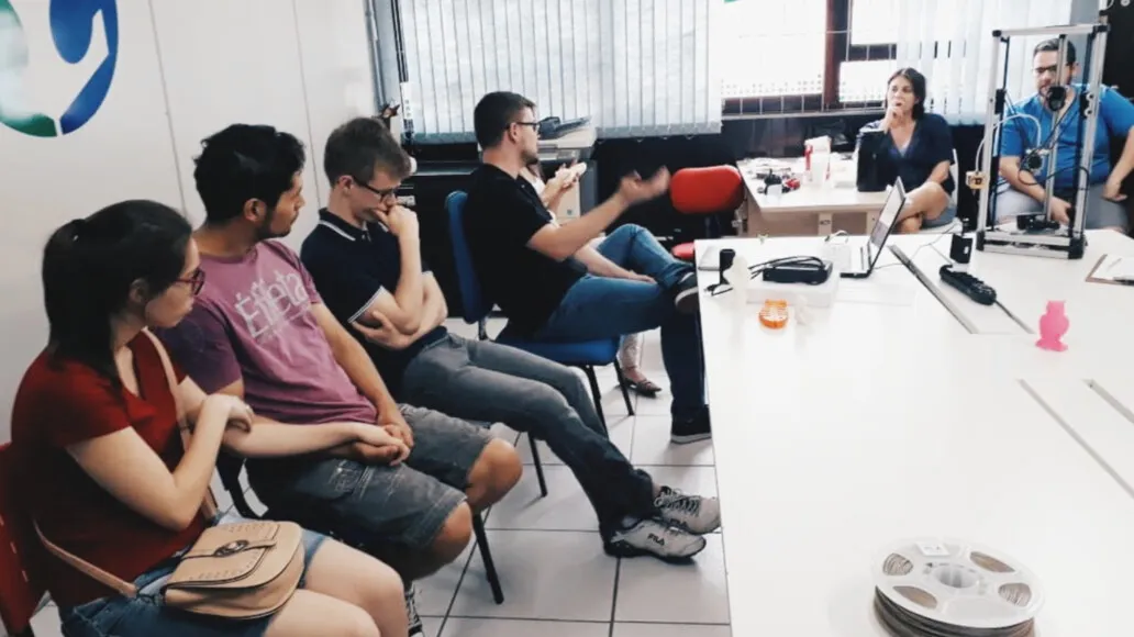 Participantes sentados acompanham uma conversa sobre o projeto Archeotech em uma sala com impressoras 3D.