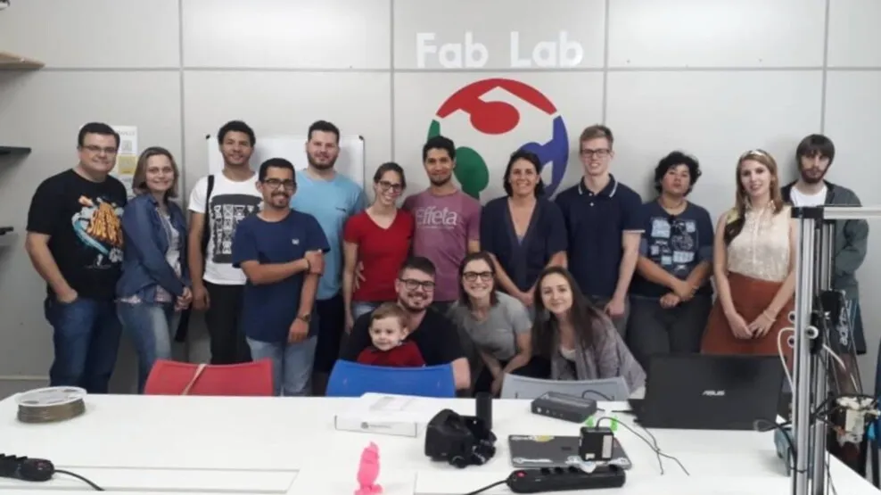 Grupo do projeto Archeotech reunido para foto no Fab Lab Joinville, diante da parede com o logotipo do laboratório.