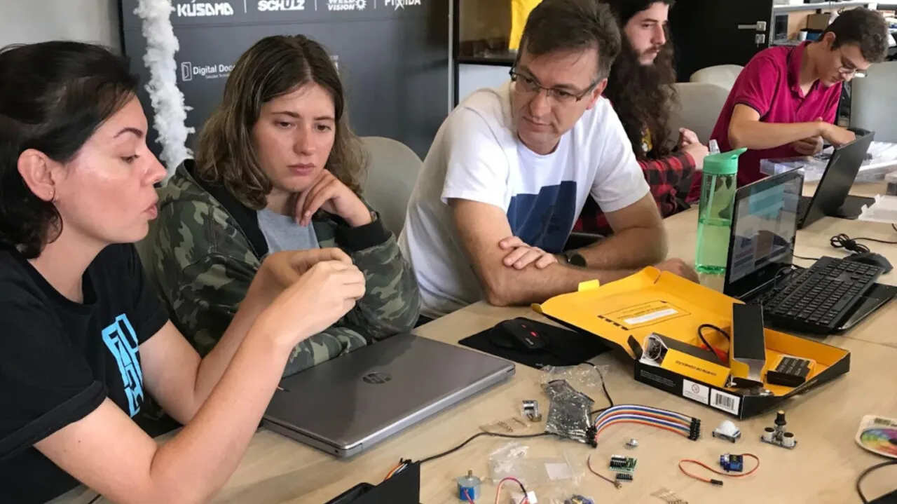 Participantes do Arduino Day montam circuitos em uma mesa com notebooks, kits de componentes e caixas de Arduino.
