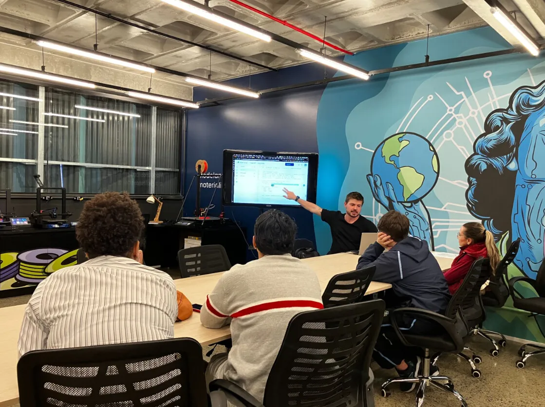 Aula no Fab Lab Joinville: um instrutor aponta para uma tela enquanto participantes acompanham sentados à mesa, com impressoras 3D ao fundo e um mural colorido na parede.