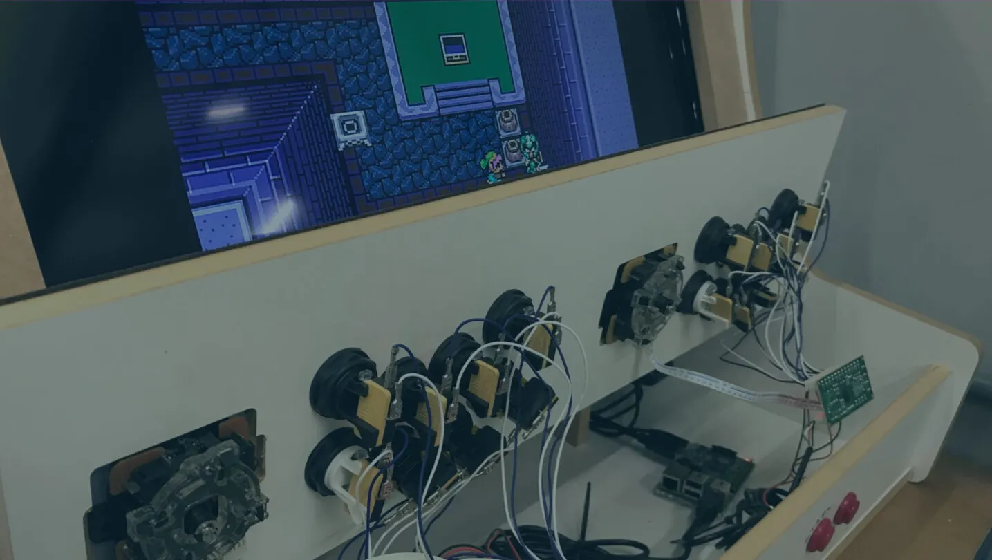Fliperama em construção no Fab Lab Joinville: painel de madeira com joysticks e botões ligados por fios, ao lado da tela com um jogo em pixel art.