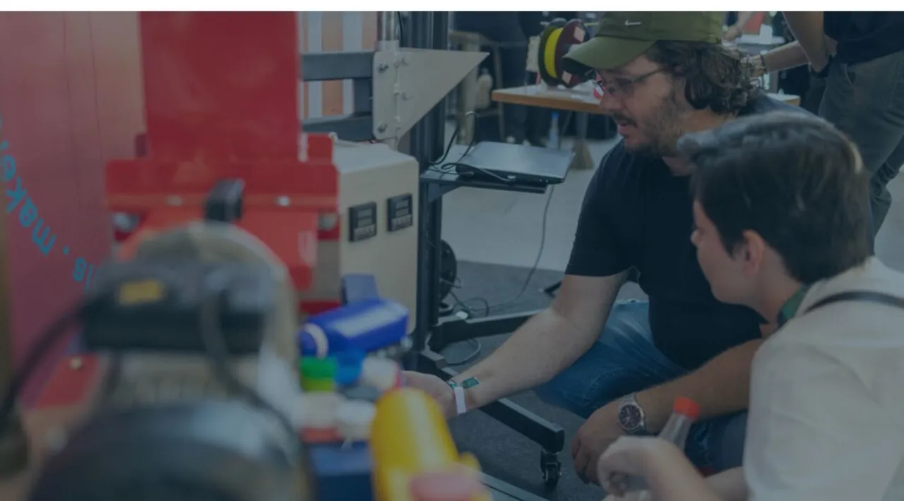Homem de boné mostra uma máquina vermelha a um jovem durante um evento maker