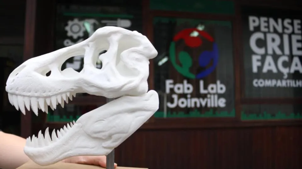 Réplica de crânio de tiranossauro impressa em 3D diante da placa do Fab Lab Joinville.