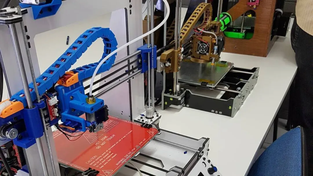 Impressora 3D de estrutura aberta, com esteira porta-cabos azul, sobre uma bancada; outra impressora aparece ao fundo.