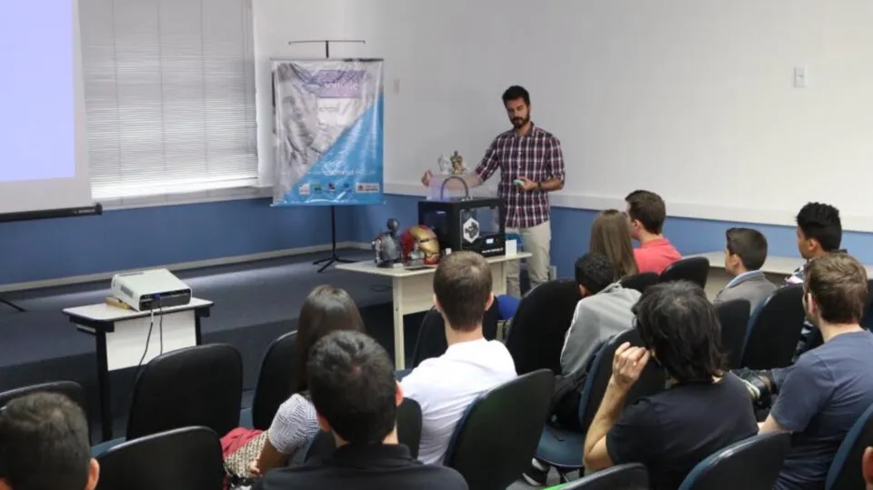 Palestrante fala ao lado de uma impressora 3D para o público sentado durante o Encontro de Impressão 3D.