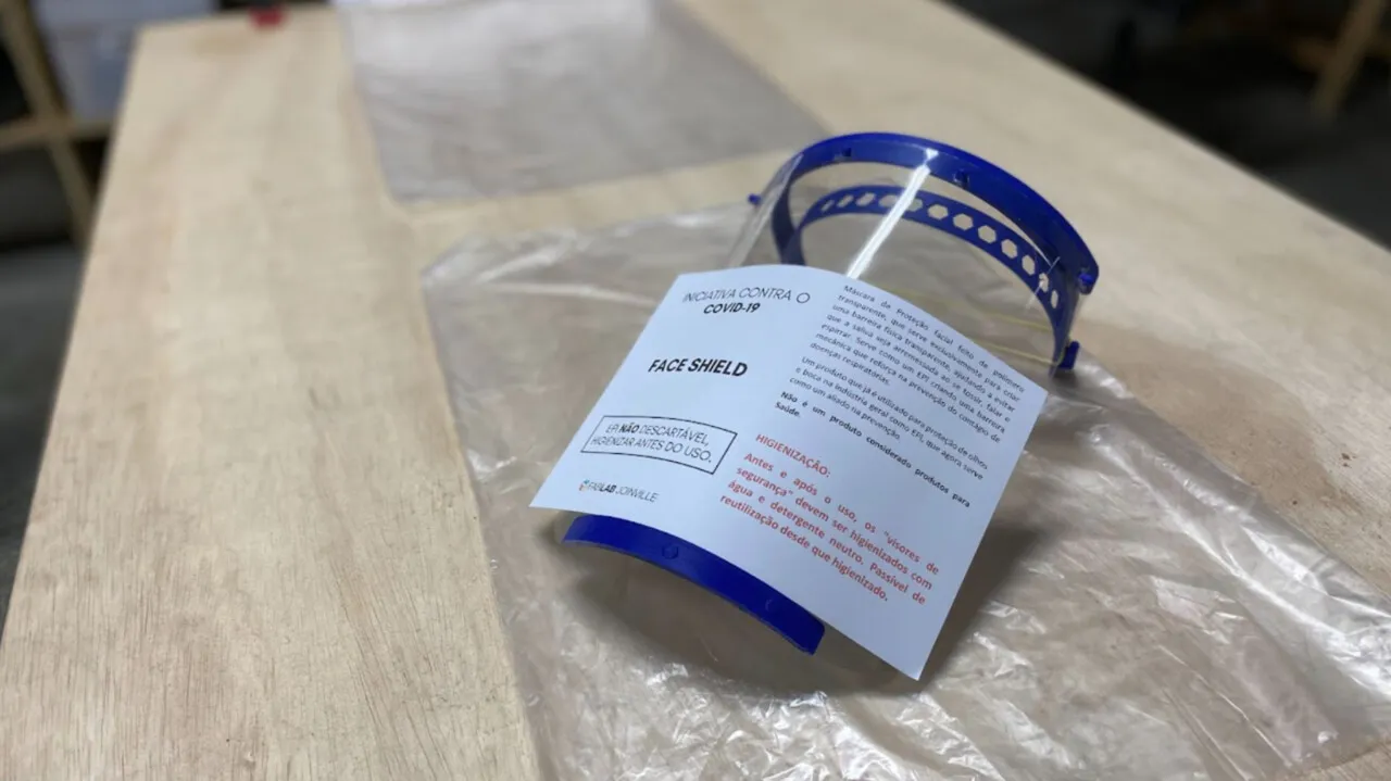 Face shield embalada em plástico, com etiqueta de instruções, sobre uma mesa de madeira.