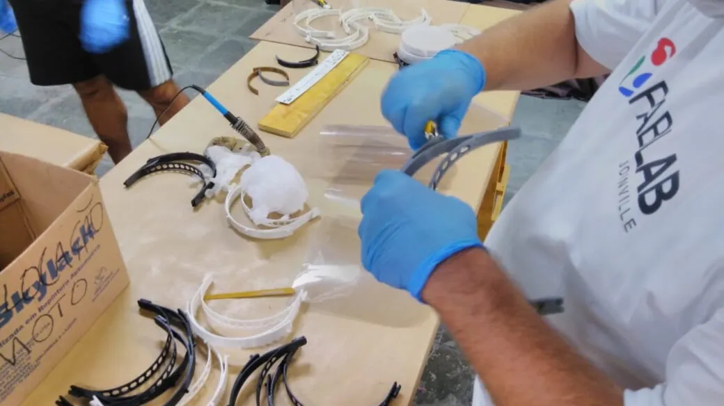 Mãos com luvas azuis montam face shields, com estruturas impressas em 3D e viseiras sobre a mesa.