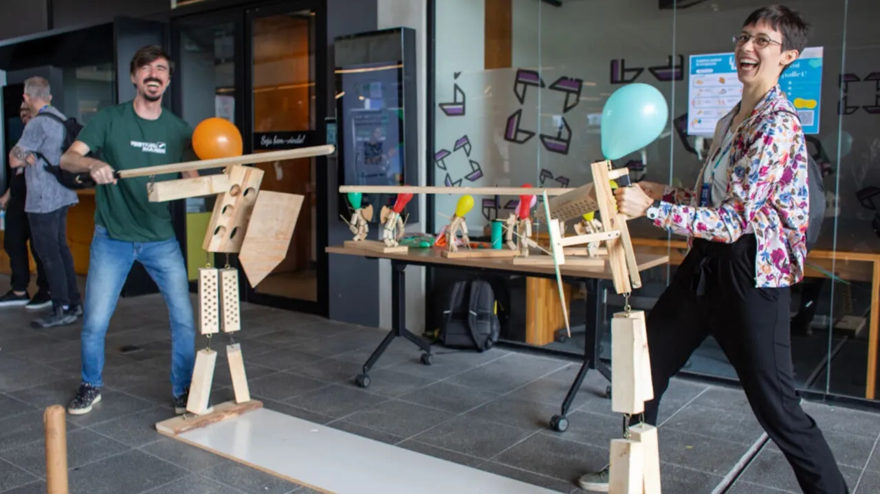 Duas pessoas sorriem enquanto movimentam grandes bonecos articulados de madeira que seguram balões no Festival Maker.