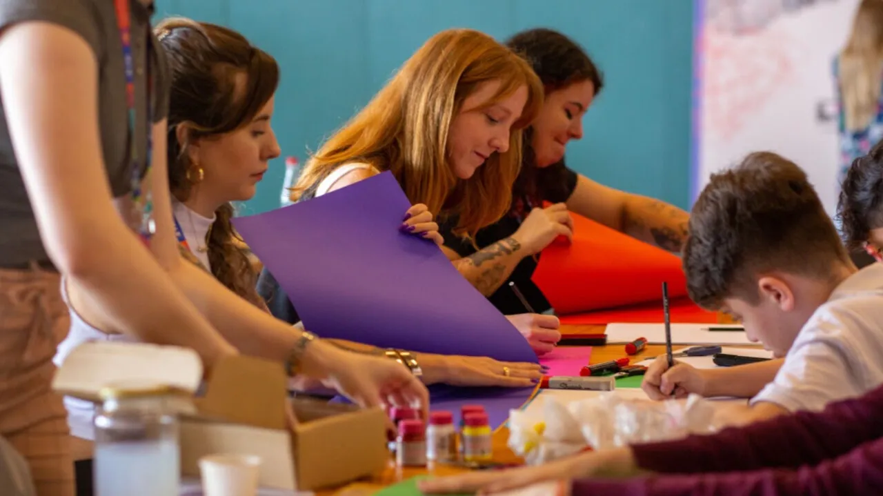 Crianças e adultos recortam papéis coloridos em uma mesa de oficina do Festival Maker.