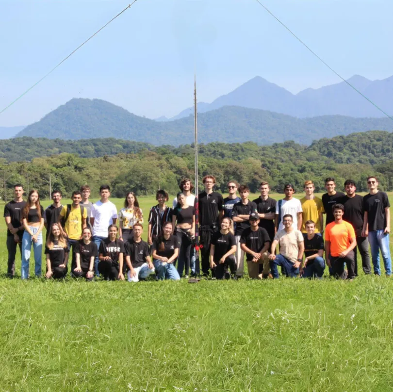 Integrantes da equipe Kosmos reunidos em um campo gramado, com montanhas ao fundo e uma torre de lançamento de foguetes.