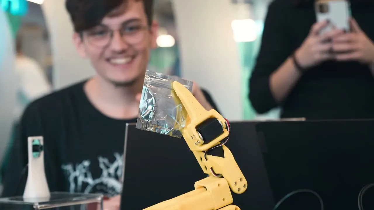 Participante sorri ao lado de um braço robótico SO-101 amarelo durante o LeRobot Arm Hackathon em Joinville