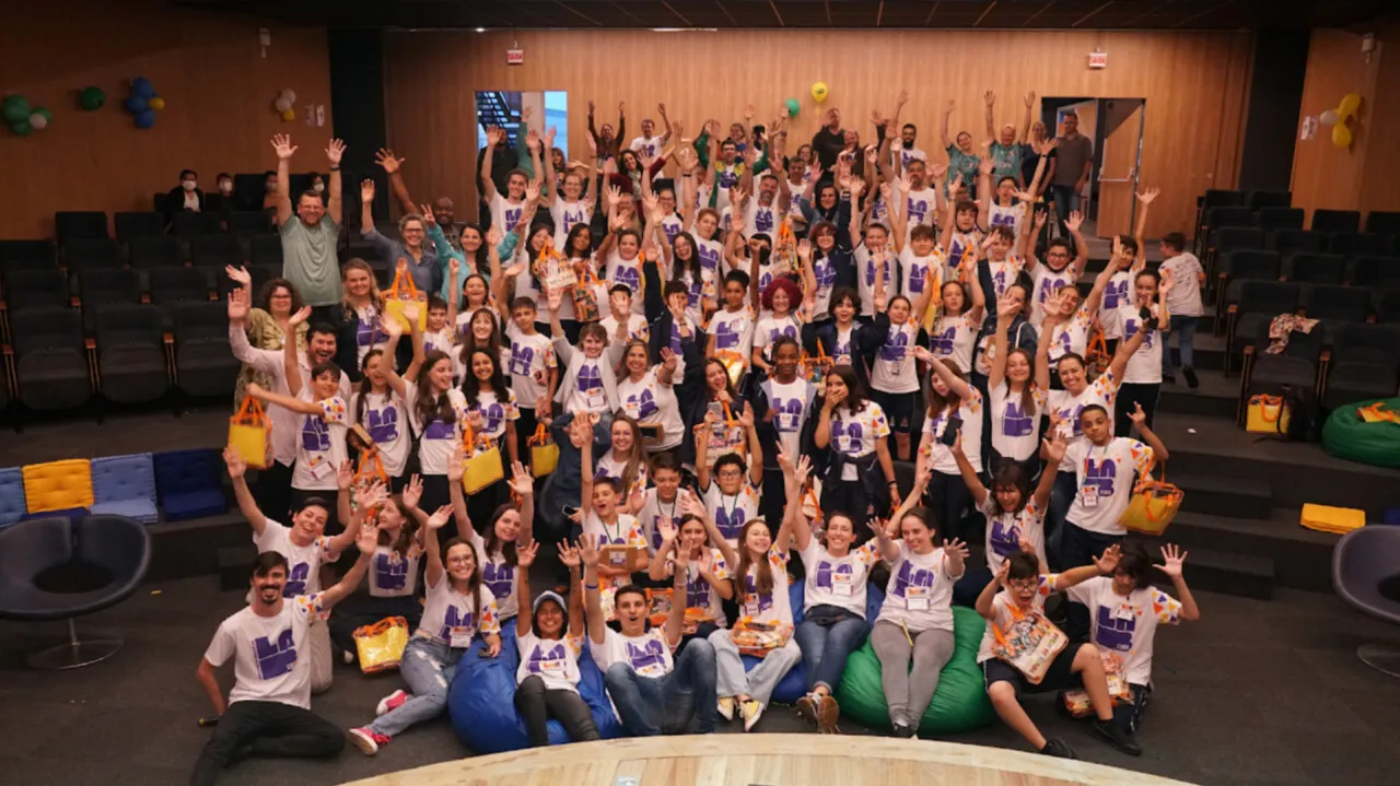 Foto oficial da Maratona Lab Code: dezenas de estudantes com camisetas do evento acenam em um auditório, sentados em pufes e nas escadas.