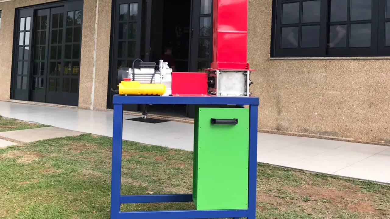 Máquina picotadora de plástico do Precious Plastic, com funil vermelho e estrutura azul e verde, montada ao ar livre.