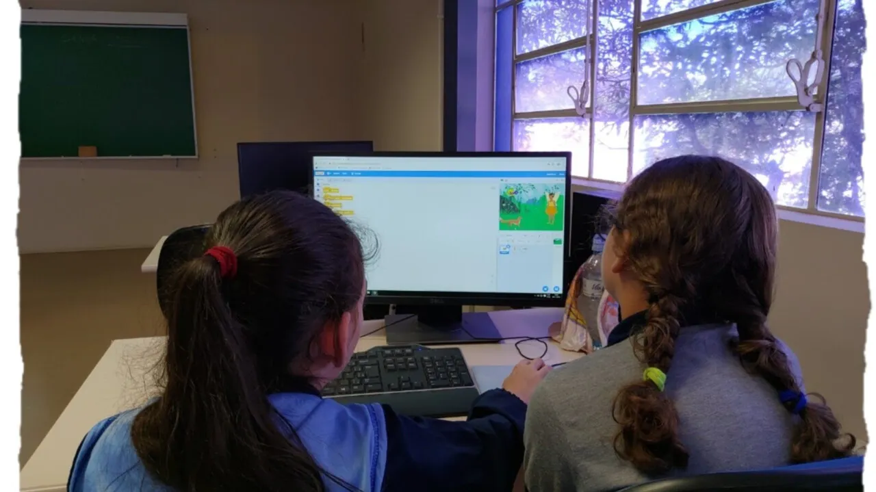 Duas alunas programam em blocos no Scratch diante de um computador em uma sala de aula.