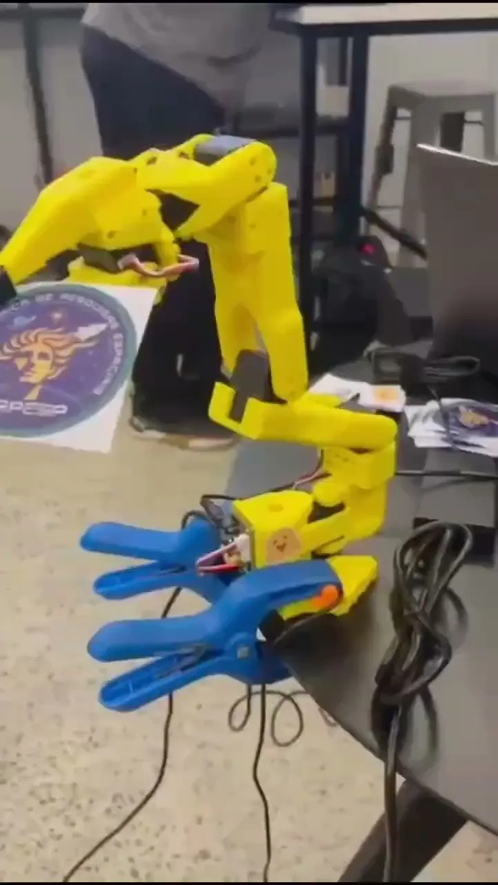 Braço robótico SO-101 amarelo com garra azul sobre a bancada, no vídeo da equipe LAPESP