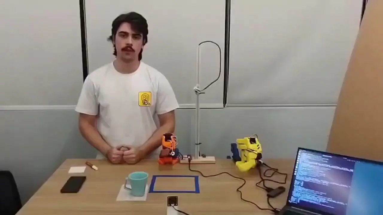 Integrante da equipe NEXA apresenta o projeto em uma mesa com dois braços robóticos SO-101 e um notebook